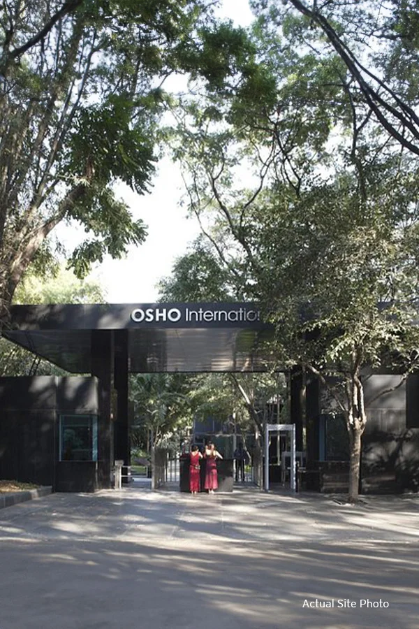 Entrance of Osho International Meditation Resort with lush greenery and visitors at the gate, under a clear sky.​